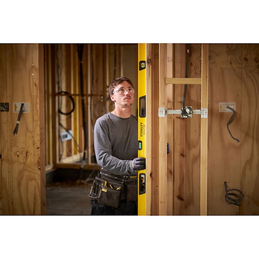 A person wearing work gloves and a tool belt is using a yellow STANLEY spirit level to check the alignment of a wooden frame at a construction site. The construction area is unfinished, with visible wooden studs and electrical wiring.