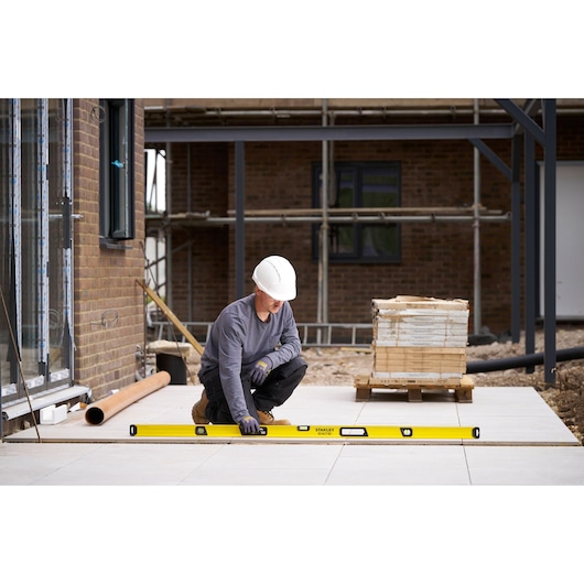 A construction worker wearing a white helmet and gloves is using a yellow STANLEY spirit level to check the alignment of outdoor pavement tiles on a building site.