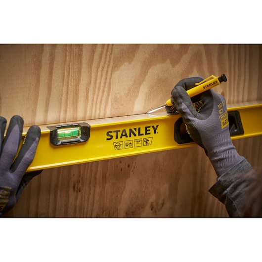 A person wearing gloves is using a yellow STANLEY spirit level and a yellow tool to mark a straight line on a wooden surface.