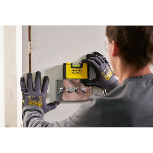 A person wearing Stanley branded gloves uses a yellow STANLEY spirit level tool (model STHT41704-0) to check alignment above a set of wall switches.