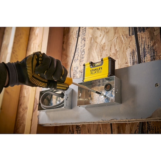 A gloved hand using a screwdriver to install or adjust a metal electrical box. A yellow STANLEY tool container is positioned on top of the box. The background shows a wooden wall under construction.