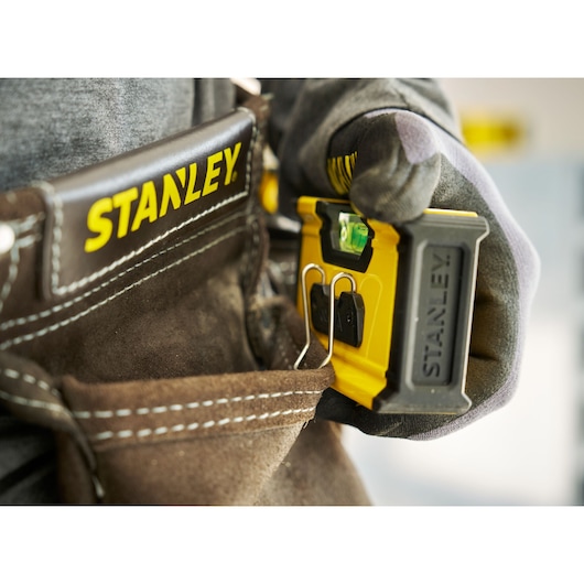 A close-up of a gloved hand placing a Stanley pocket level into a leather tool belt with the Stanley logo visible.
