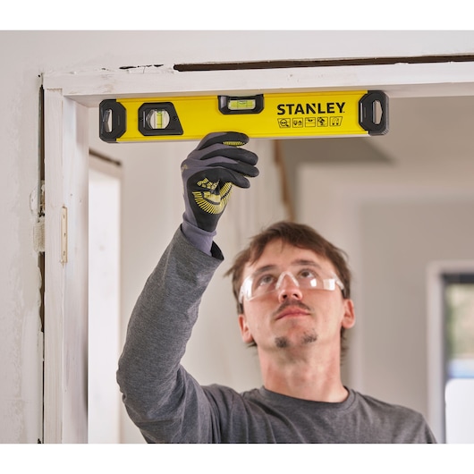 A person wearing work gloves uses a yellow STANLEY spirit level to check the alignment of a door frame.