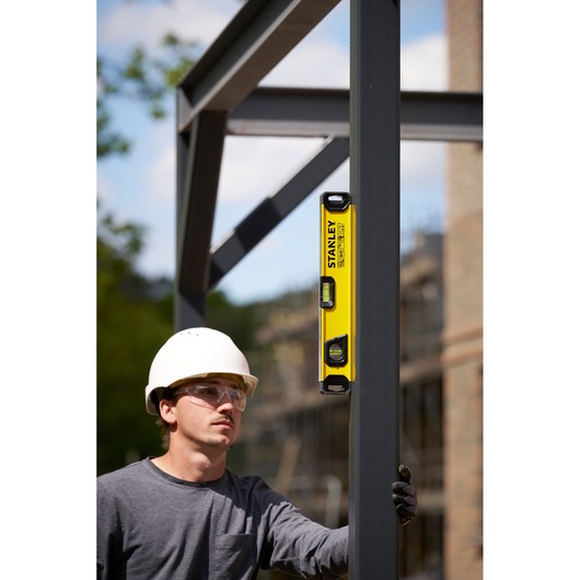 A person wearing a white safety helmet and grey shirt uses a yellow STANLEY spirit level (model STHT41716-0) to check the vertical alignment of a metal beam at an outdoor construction site. The person's face is blurred.