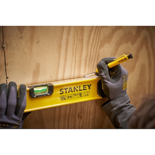 A person wearing gloves uses a Stanley spirit level and a marking tool on a wooden surface. The Stanley level is bright yellow with black markings and a green bubble vial.