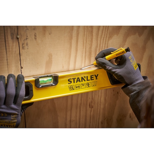A person wearing work gloves uses a Stanley yellow spirit level and marking tool to draw a straight line on a wooden surface.