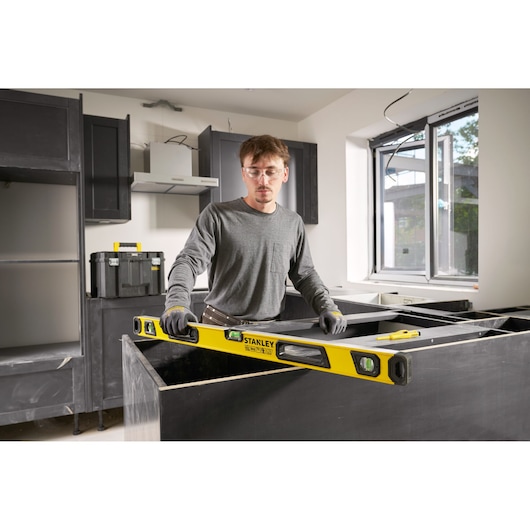 Person using a yellow STANLEY spirit level (model STHT41736-0) in a kitchen under construction. The individual is wearing gloves and grey clothing, aligning a countertop. Construction tools and materials are visible in the background.