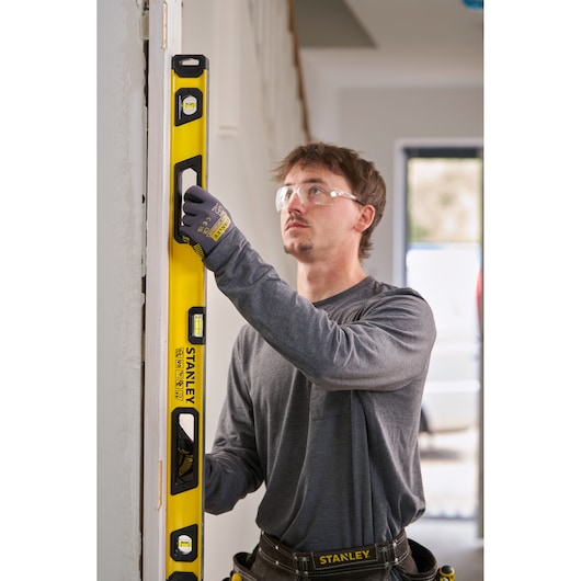 A person wearing work gloves and a grey shirt uses a yellow Stanley spirit level to check the vertical alignment of a door frame inside a building. The Stanley branding and measurement markings are visible on the tool.