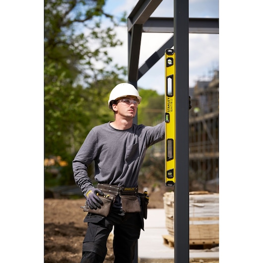 A construction worker using a yellow STANLEY spirit level (SKU: STHT41736-0) to check the alignment of a vertical metal beam at an outdoor construction site. The worker is wearing a hard hat, gray shirt, gloves, and a tool belt. The background features trees and construction materials.