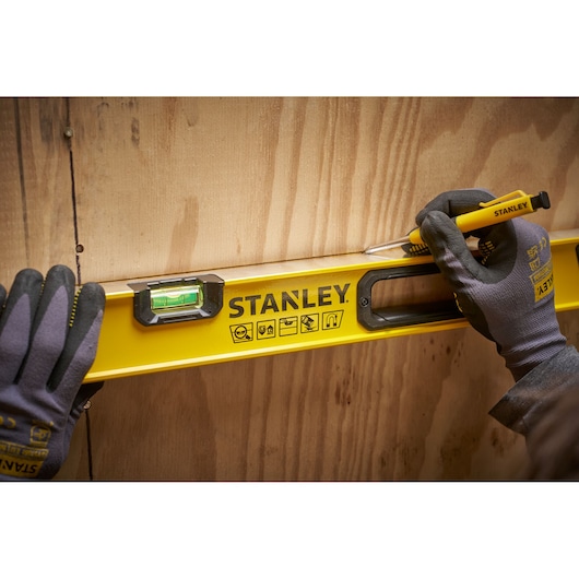 A person wearing gloves is using a yellow STANLEY spirit level against a wooden surface, marking a line with a STANLEY utility knife.