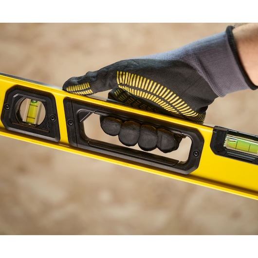 A close-up of a gloved hand holding a yellow Stanley spirit level with black grip, showing the bubble vials clearly.