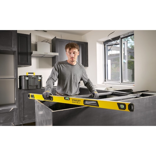 Person using a Stanley spirit level to measure and align a countertop in a kitchen under construction, with toolboxes and cabinets visible in the background.