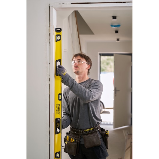 A person wearing work gloves and a tool belt is using a yellow STANLEY spirit level to check the vertical alignment of a door frame in an indoor construction setting. The person's face is blurred.