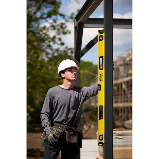 A construction worker wearing a white hard hat and a tool belt is using a yellow STANLEY spirit level against a metal frame at an outdoor construction site.