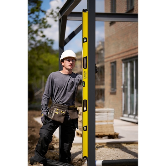 A person wearing a white safety helmet and a tool belt uses a yellow STANLEY spirit level to check the vertical alignment of a metal beam at an outdoor construction site.