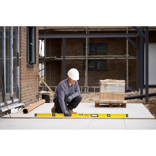 A construction worker wearing a white hard hat and gloves is using a yellow STANLEY spirit level to check the alignment of outdoor paving tiles at a building site.