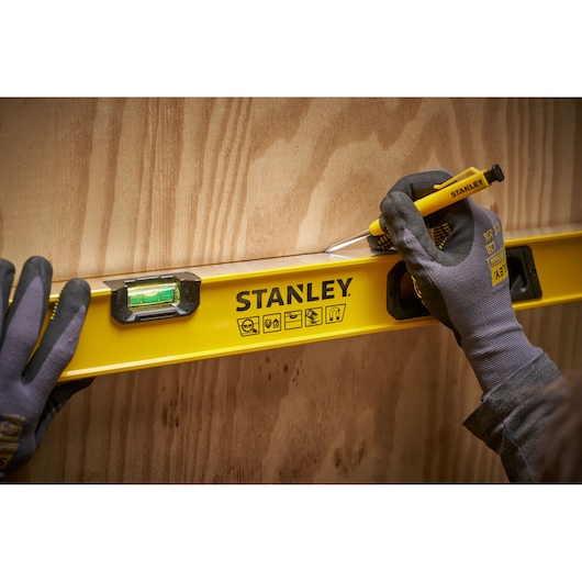 A person wearing work gloves uses a yellow STANLEY spirit level against a wooden surface, marking a line with a STANLEY tool.