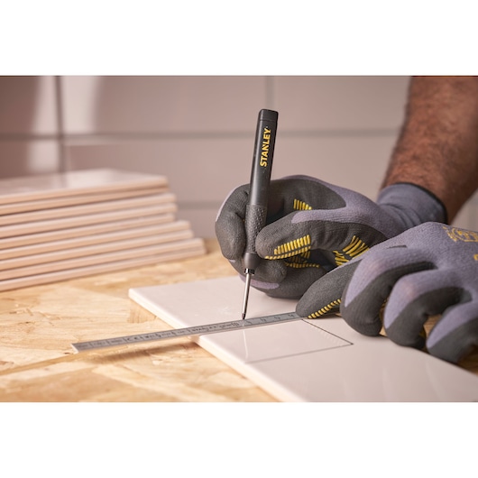 Gloved hands use a STANLEY marking tool and ruler to draw lines on a tile for precise measurement.