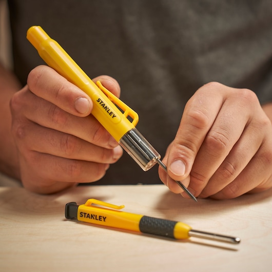Person using a yellow STANLEY marking tool on a table. Another similar tool lies beside it.